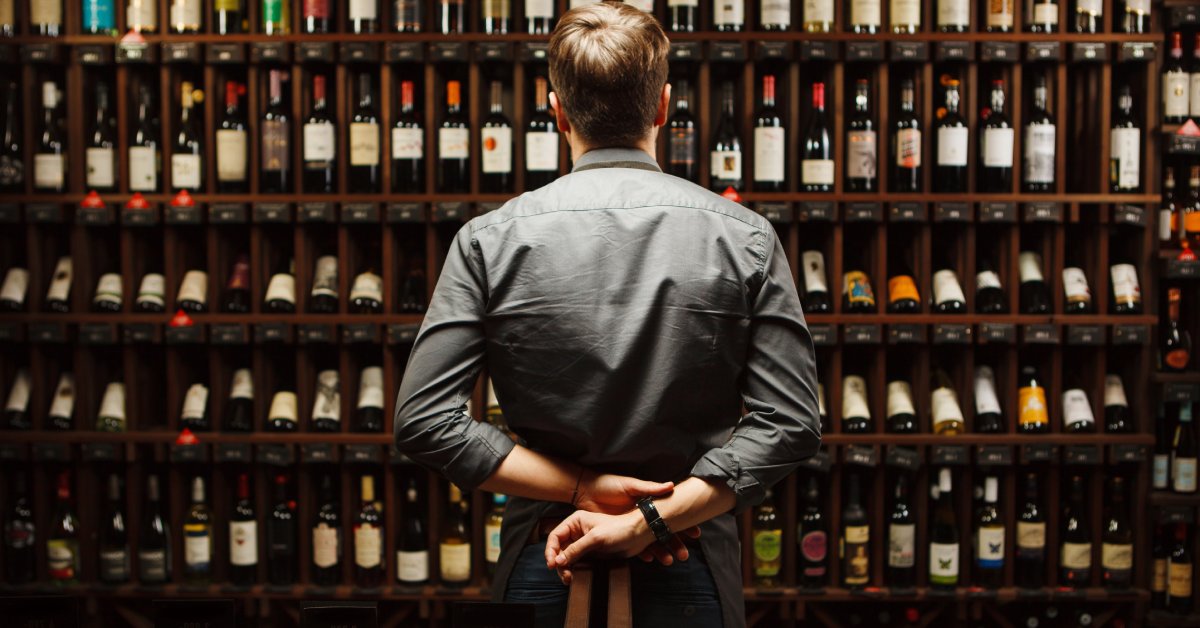A man wearing a nice gray shirt stands with his hands behind his back, staring at a wall of wine bottles.