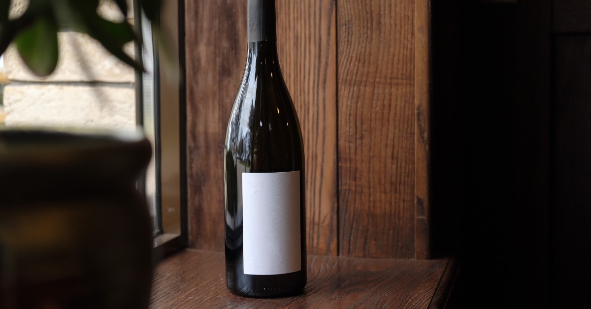A dark-colored wine bottle sits on a wooden surface with a blank, white label on the front. A window is next to the wine.