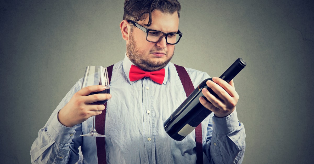 A man wearing a button-down shirt and a red bow tie holds a glass of red wine in one hand and the bottle in the other.
