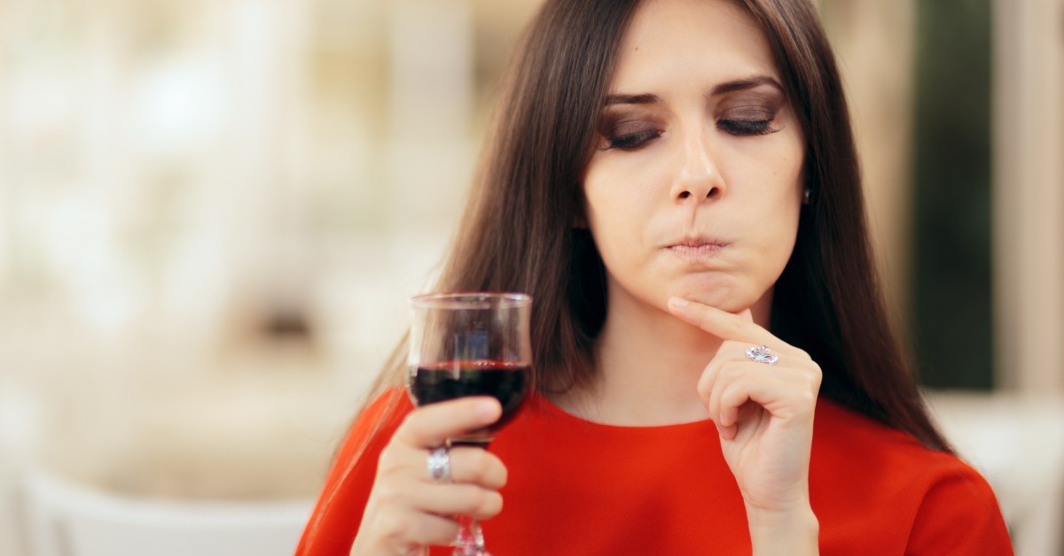 A woman in a red dress holds a clear wine glass filled with red wine. She looks at the glass with a puzzled look.