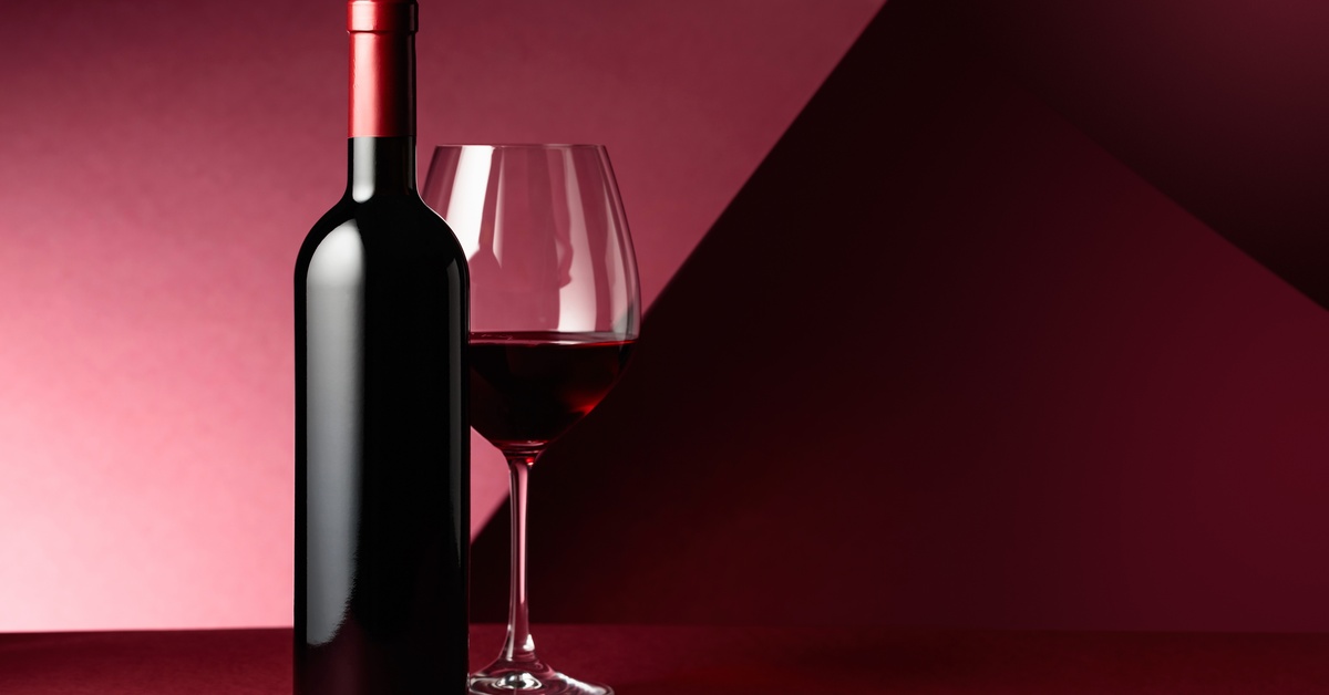 An unopened bottle of red wine sits next to a wine glass halfway full of red wine. The background is burgundy.