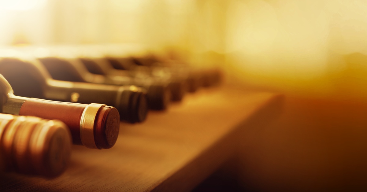 A row of unopened wine bottles lay on a wooden shelf on their sides. The sun shines on the bottles.