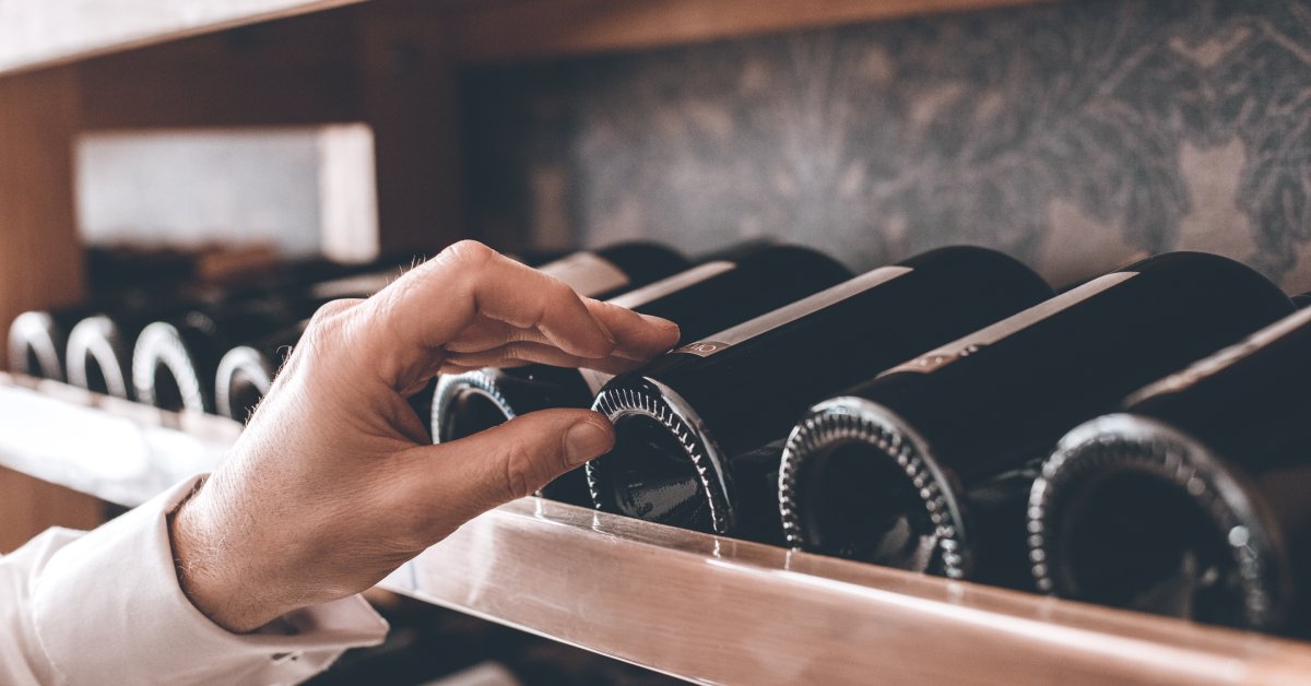 A hand reaches toward several bottles of wine. The bottles are placed onto a wooden display rack.