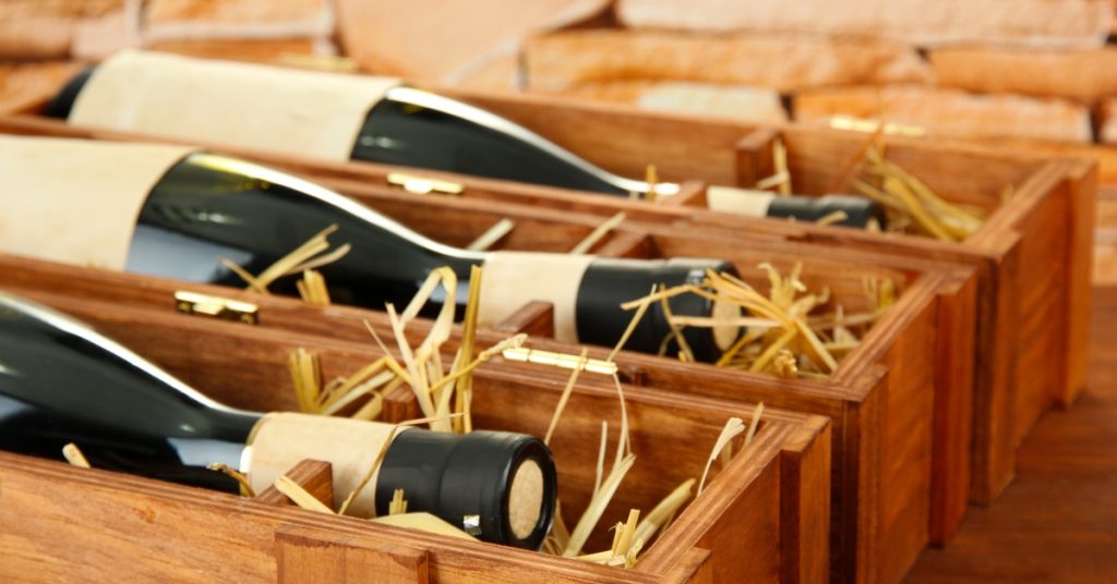 Bottles of wine with beige-colored labels on them lay on top of straw in thin wooden boxes on a wood surface.