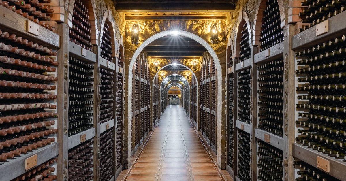 A long wine cellar with stacks of horizontal wine bottles stacked on shelves. The floor of the cellar is brown tile.
