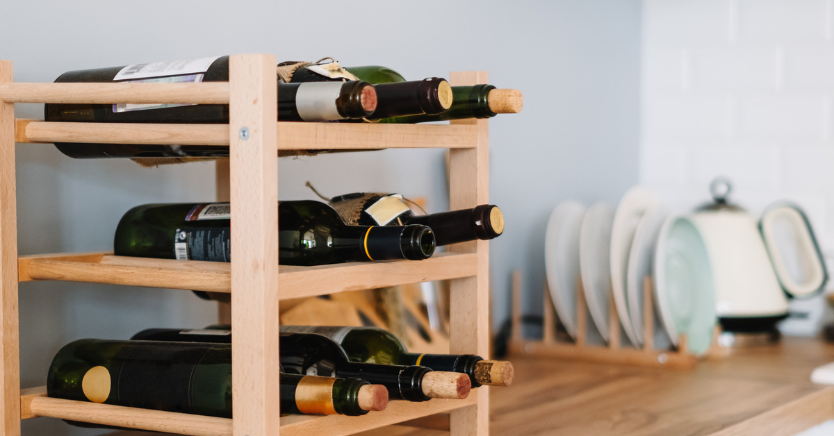 A small, wooden wine rack with eight wine bottles on it sitting on a kitchen counter near some dishes.
