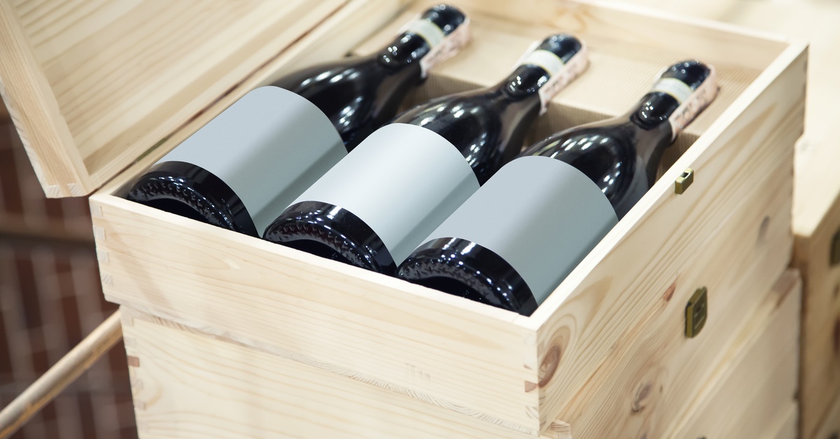 Three wine bottles with gray, blank labels lie horizontally in a light wooden box with the lid open.