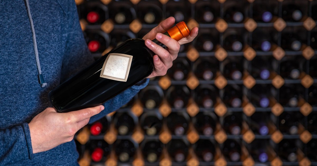 A person in a blue hoodie holds a wine bottle in front of a large wine rack full of many other bottles of wine.