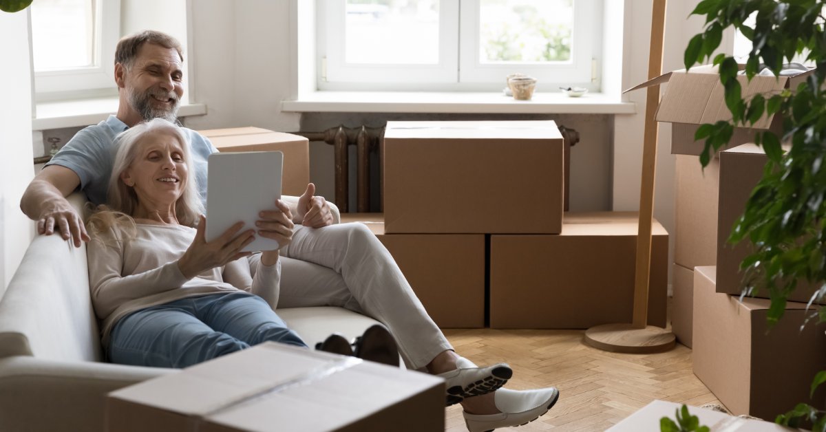 An older man and woman relax on a couch, using an ipad and smiling, while surrounded by moving boxes.