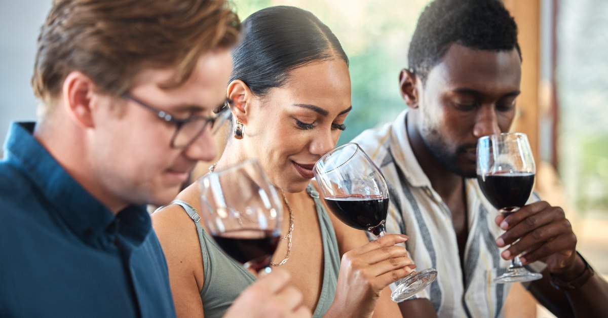 Three young adults sniff glasses of red wine at an indoor wine tasting event on a sunny day.