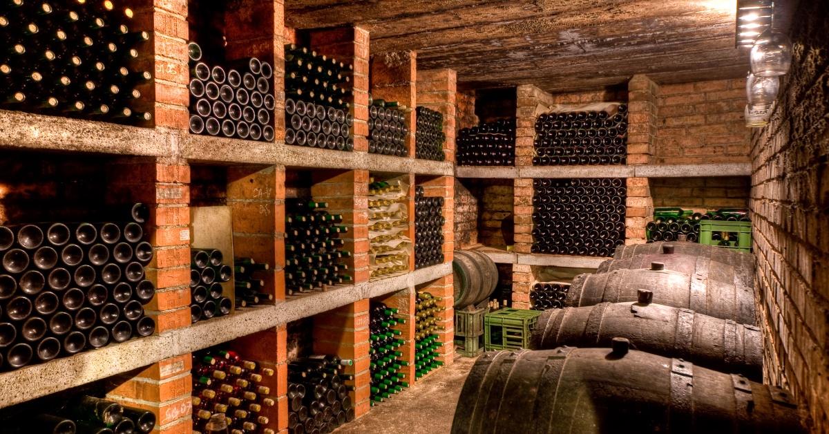 Hundreds of corked wine bottles stored horizontally on wooden shelves in a winery’s brick wine cellar.