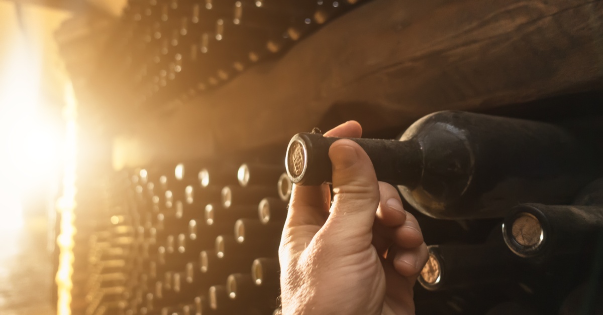 A hand grabs the neck of a wine bottle to pull it off the shelf of a winery’s wine cellar as the sun goes down.