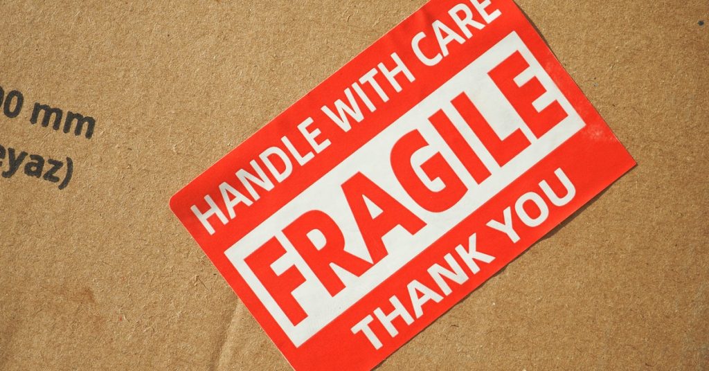 A close-up of a red and white sticker that says “handle with care fragile thank you." The sticker is on a cardboard box.