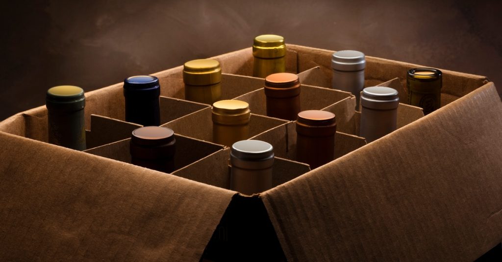 Twelve wine bottles with various colored seals over their corks set up in a cardboard box and separated by a cardboard divider.
