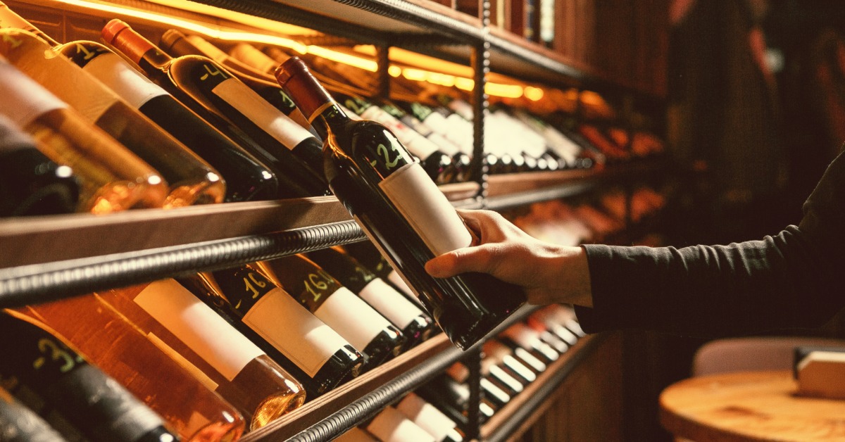 Someone stands in the wine cellar holding a bottle of wine as they read the label. The cellar is dark and full of inventory.