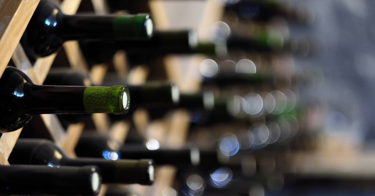 A close-up of wine bottles arranged neatly on a wooden rack with more wine bottles going out of focus in the distance.