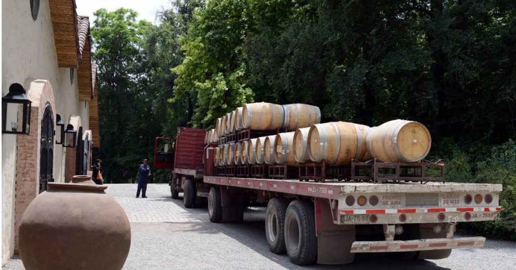  A semi truck with a long trailer loaded up with dozens of wine barrels outside of a winery with the truck driver standing outside.