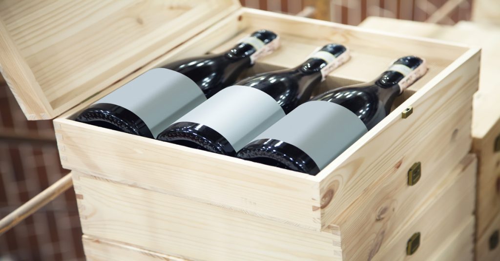 Three bottles of red wine packaged in a wooden box with plain white labels stacked on top of other wooden boxes.