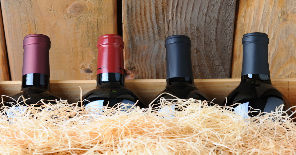 A close-up on four wine bottles with dark red and black wrappers resting in a wooden box and surrounded by fluffy hay packaging.