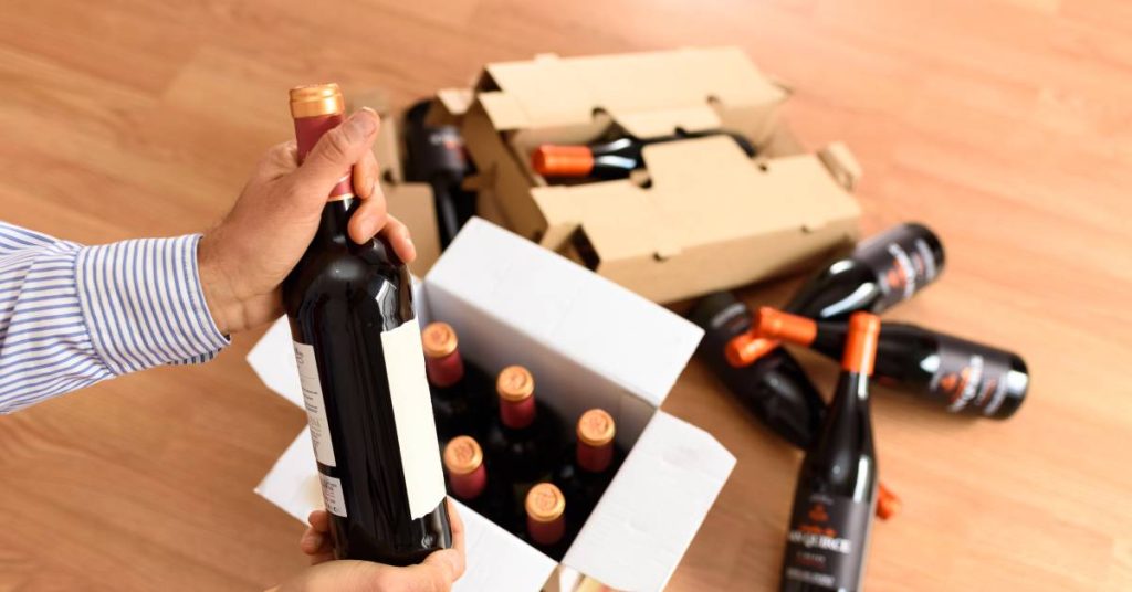 Multiple bottles of red wine sent by courier in cardboard boxes, one bottle of wine held up by a man in a blue and white striped shirt.