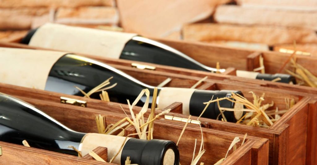 Three bottles of red wine in wooden gift boxes with hay for cushioning during transportation, on a stone wall background.