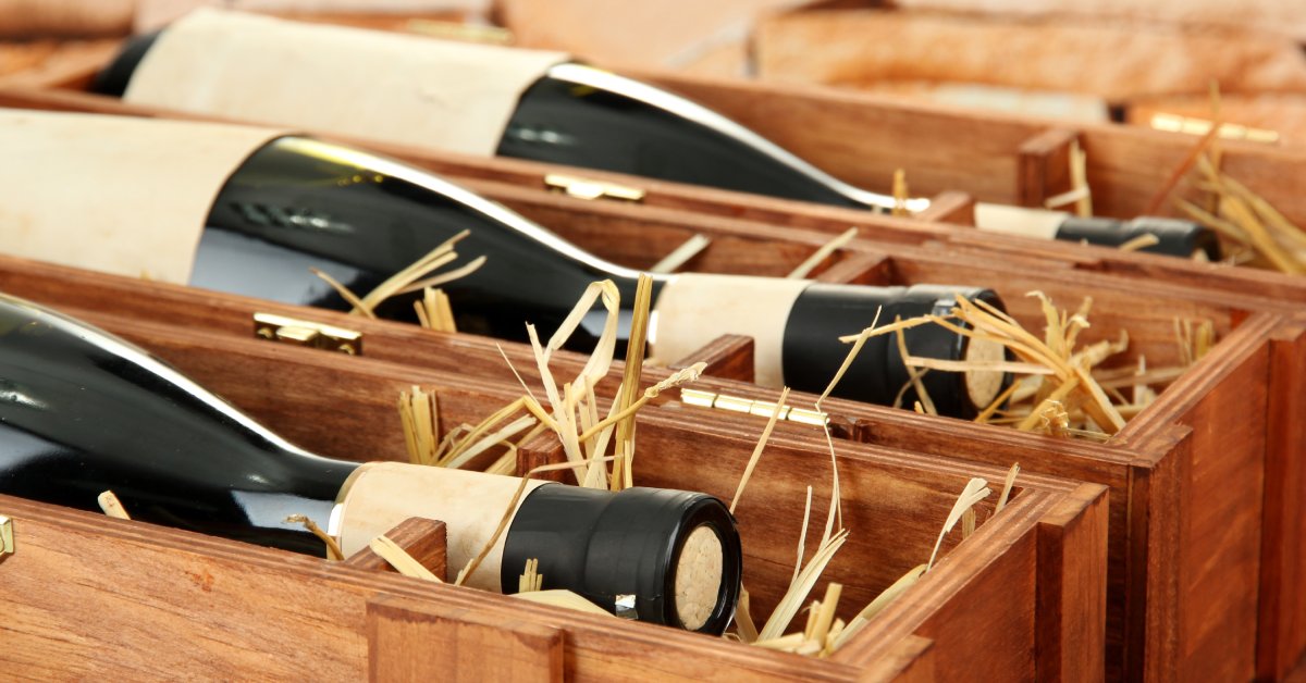 Bottles of red wine sit in a wooden crate with paper filler. The bottles are room temperature and ready for storage.