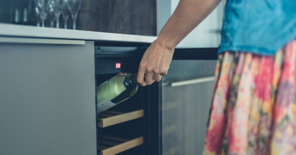 A person stands in front of their wine fridge in a blue top and floral skirt. They're pulling a white wine out of storage.